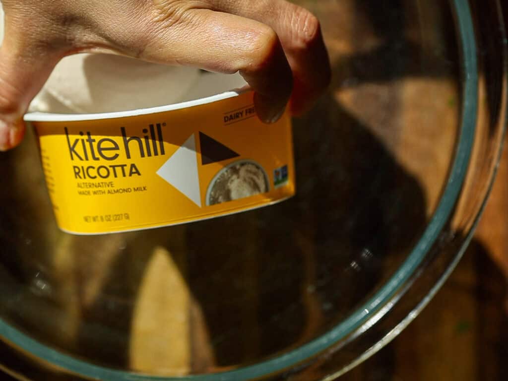 Hand holding a container of Kite Hill dairy-free ricotta made with almond milk, against a wooden background. The container is labeled as an alternative product.